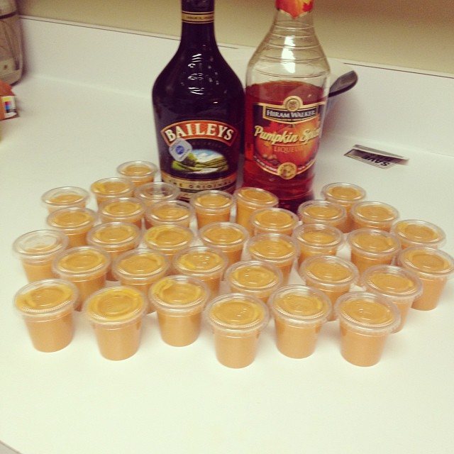 Small clear plastic cups filled with orange pumpkin pudding arranged on a white countertop near liquor bottles.