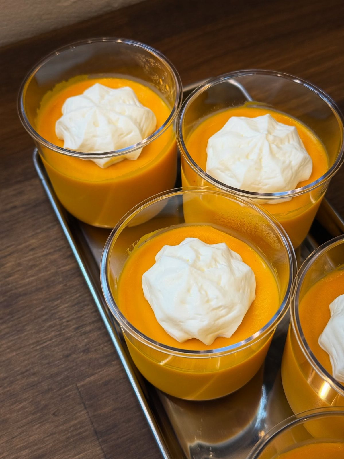 Clear plastic cups filled with orange pumpkin pudding topped with white whipped cream on a tray.