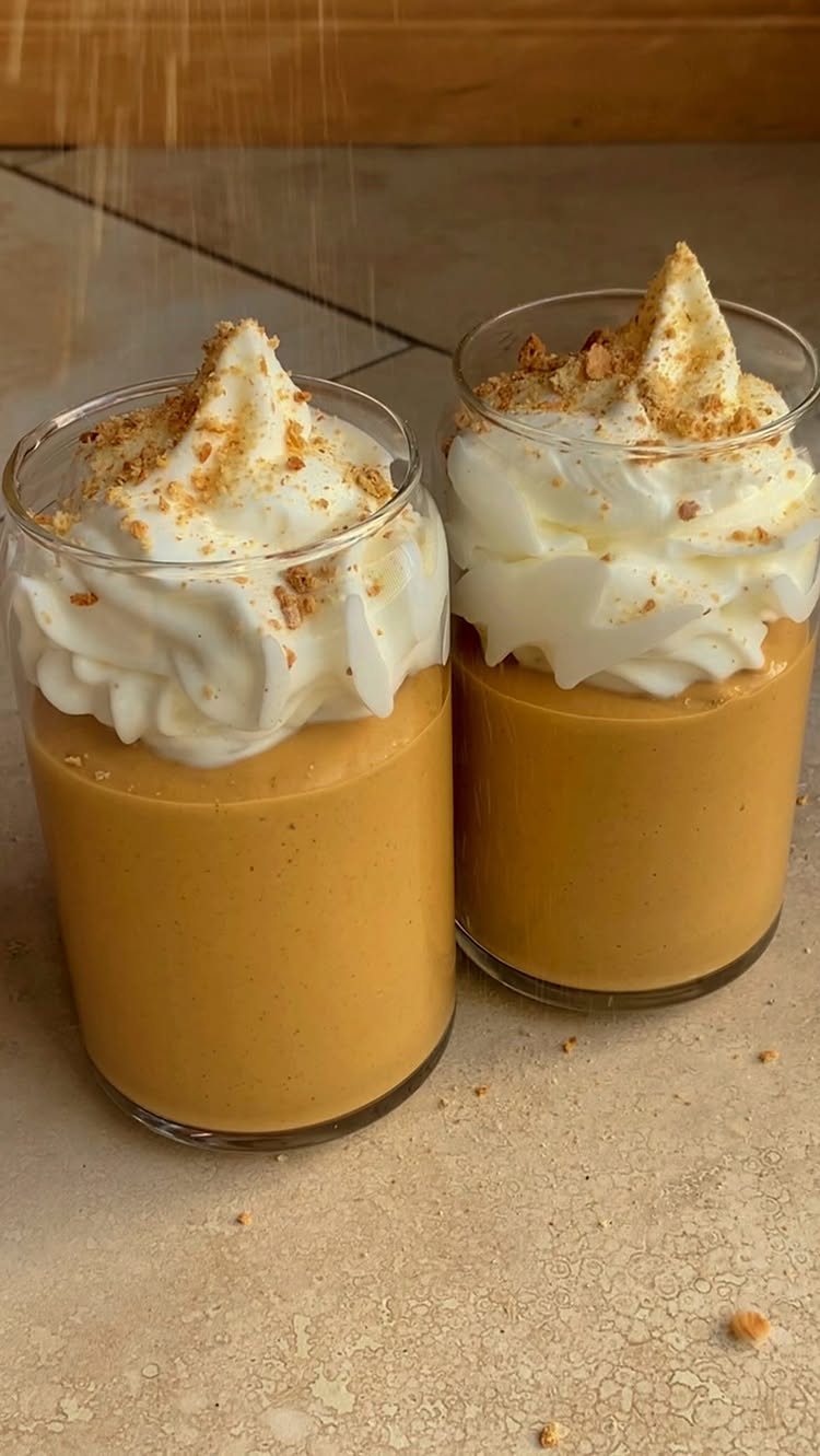 Two glasses of pumpkin pudding topped with whipped cream and graham cracker crumbs on a tan surface.