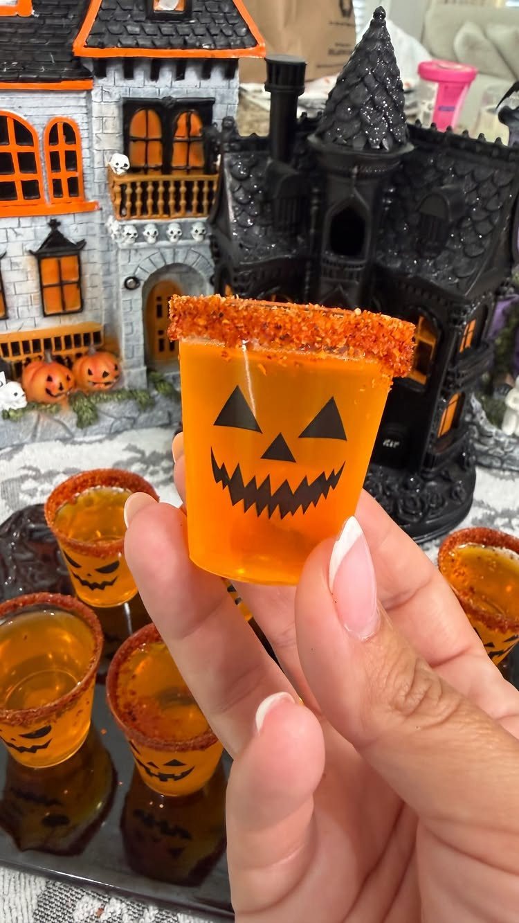 A hand holding an orange jack-o'-lantern shot glass with a sugared rim near a miniature haunted house.