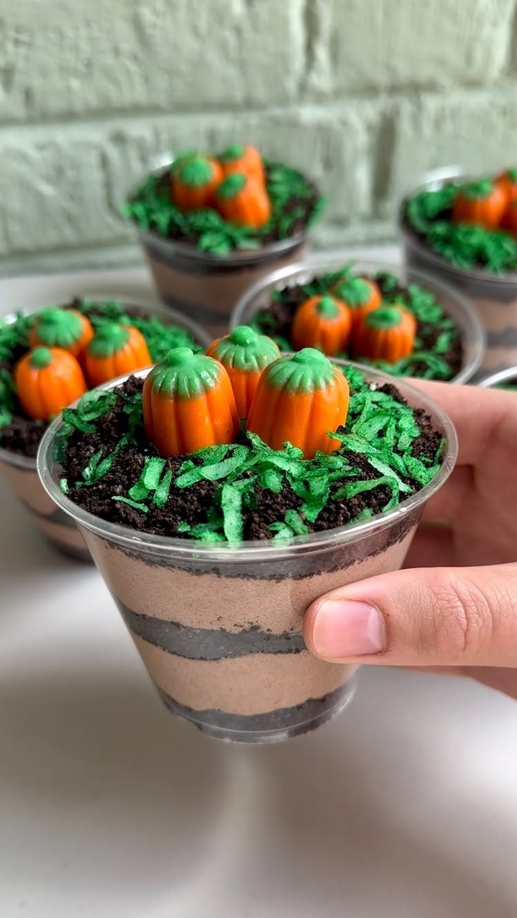 A hand holding a clear cup of chocolate pudding topped with green sprinkles and tiny orange candy carrots.