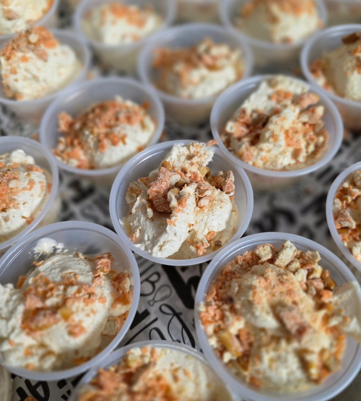 Small plastic cups filled with creamy pumpkin pudding topped with orange crumbles on a patterned surface.