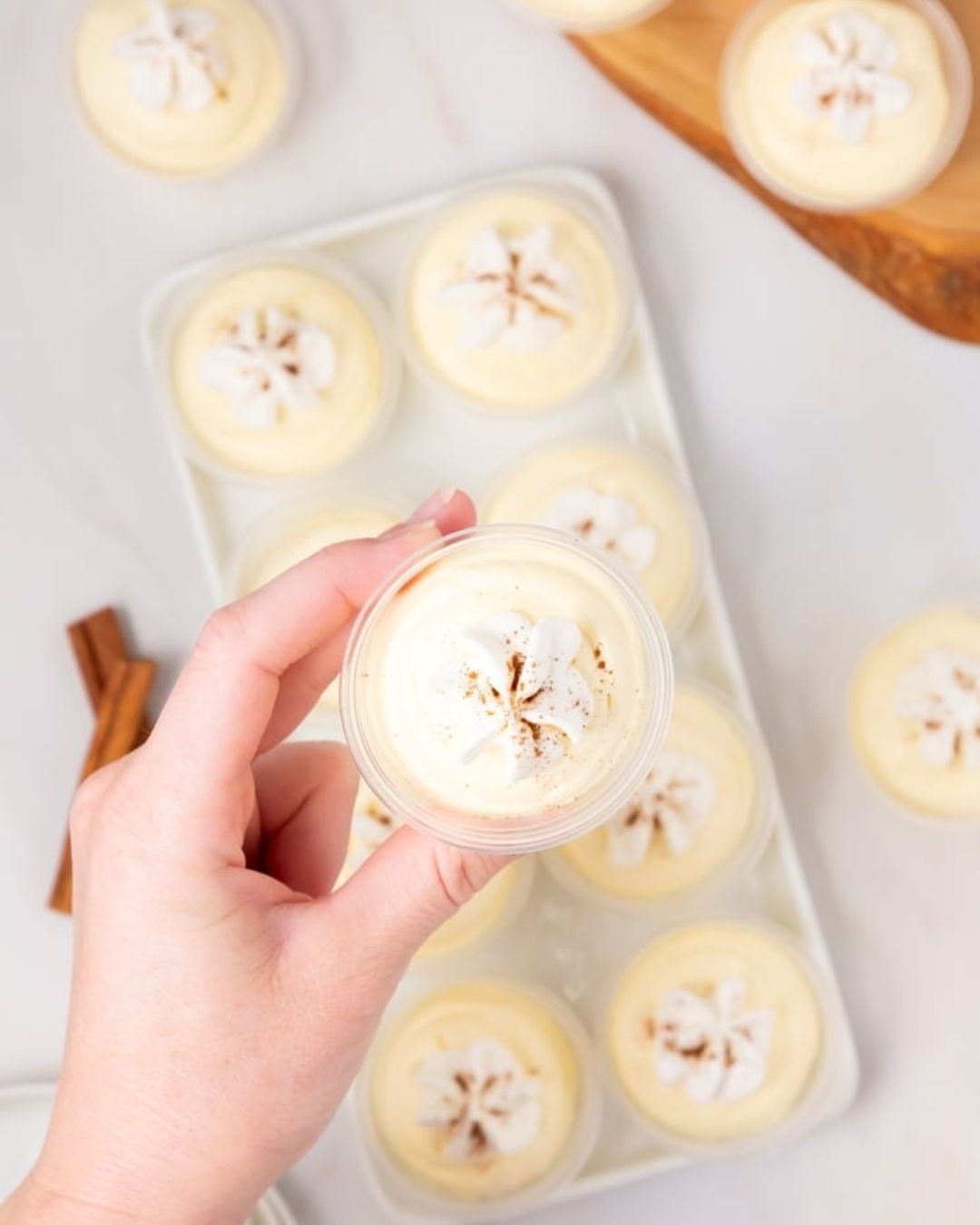 A hand holding a vanilla pudding shot glass topped with whipped cream and cinnamon sprinkles.