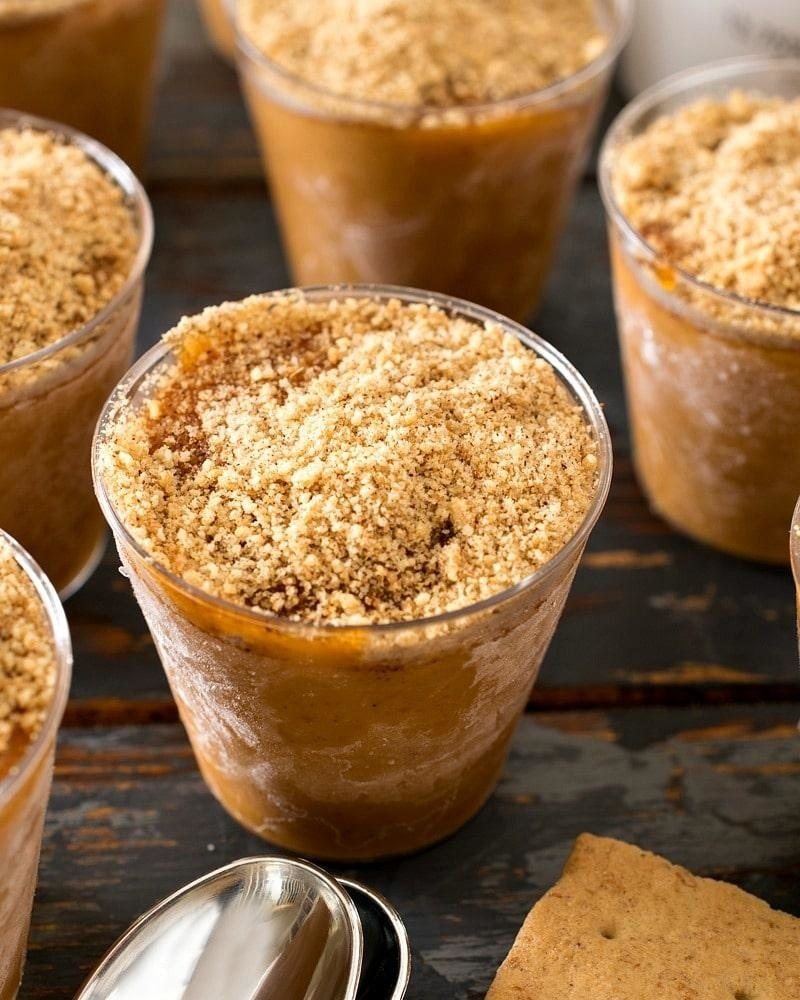 Small plastic shot glasses filled with brown pudding and golden crumble topping on wood.