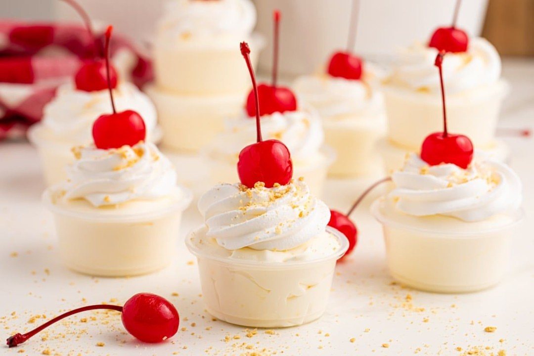 Creamy white pudding shots topped with whipped cream, graham cracker crumbs, and bright red cherries.