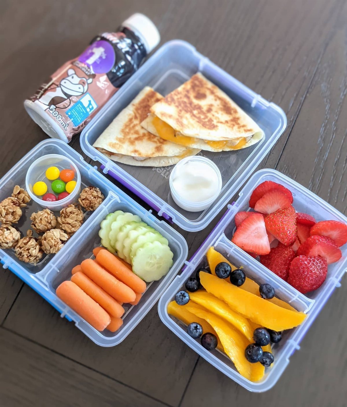 19 School Lunch Ideas for Real Mornings 1 Three bento lunch boxes containing quesadillas, vegetables, granola, and fresh fruit on a wooden table.