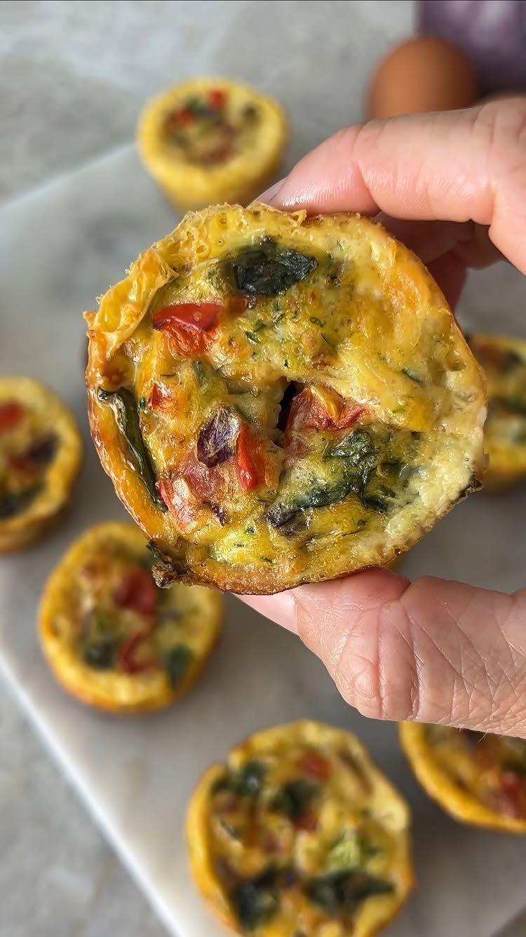 19 School Lunch Ideas for Real Mornings 7 A hand holds a savory egg bite filled with spinach and red peppers over a marble surface.