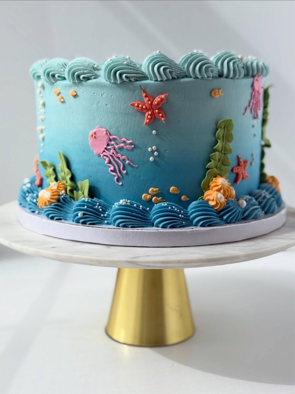 8 Undersea Cake Ideas From Fondant Reefs to Jellyfish Buttercream 7 Blue ombre cake decorated with pink jellyfish, orange starfish, and green seaweed on a marble stand.