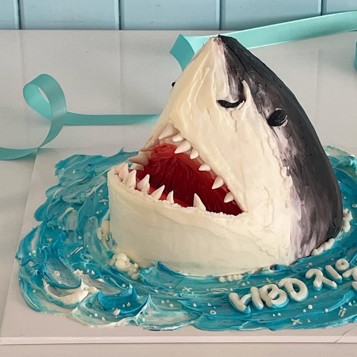8 Undersea Cake Ideas From Fondant Reefs to Jellyfish Buttercream 5 A shark head cake emerging from blue frosting waves on a white surface.