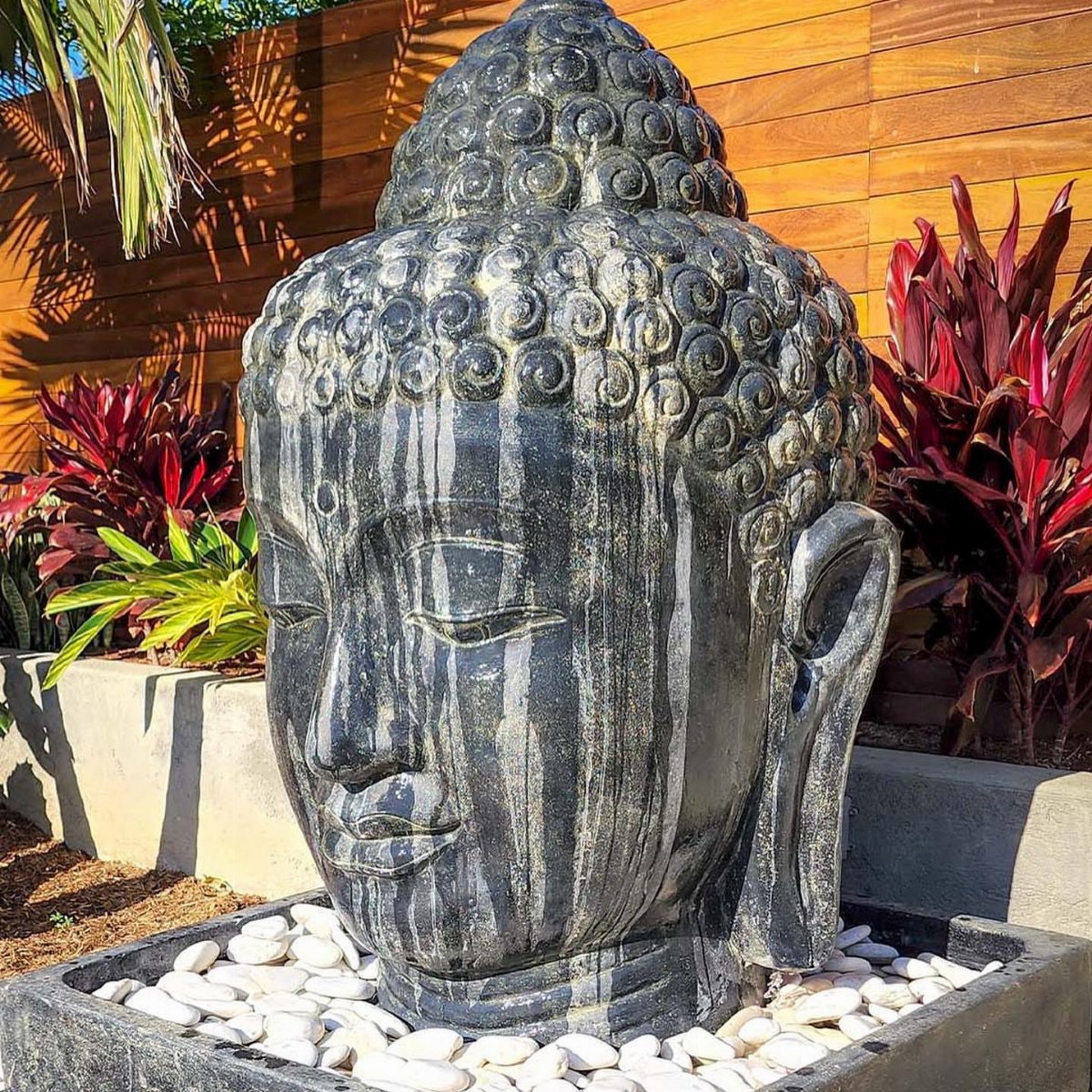10 Backyard Fountain Ideas for Patios, Walls & Small Spaces 8 A large dark stone Buddha head sculpture resting in a bed of white pebbles.