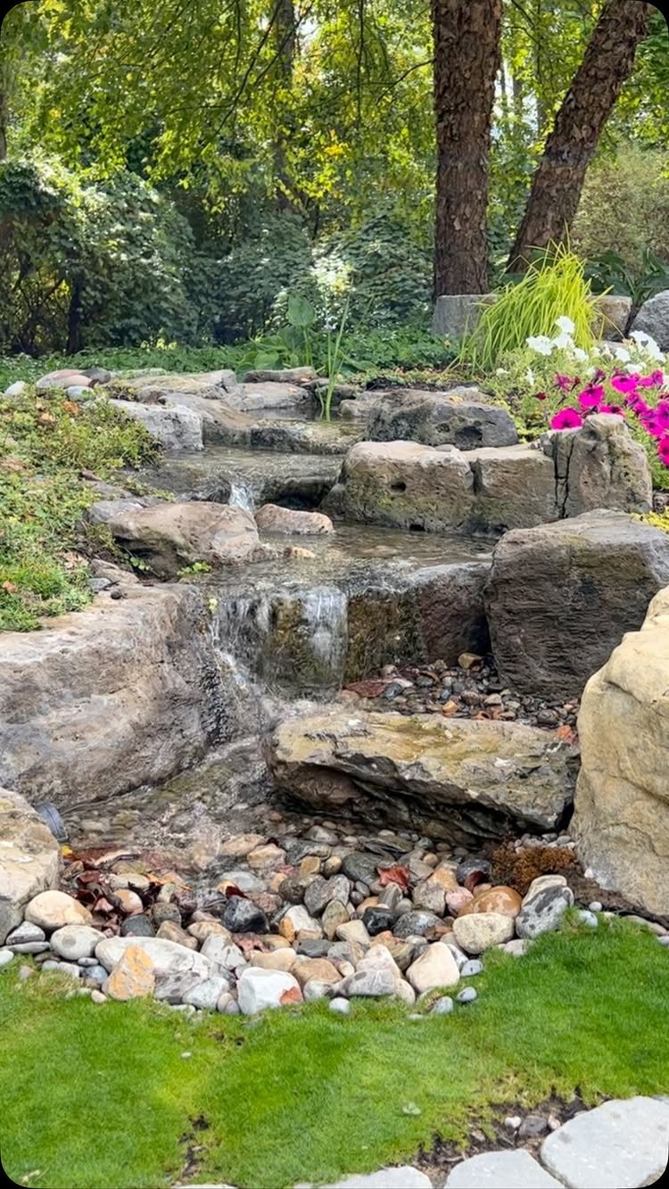10 Backyard Pond Ideas That Bring Water, Sound and Life to Your Yard 2 Water cascading over large natural rocks in a garden stream lined with pebbles and surrounded by greenery.