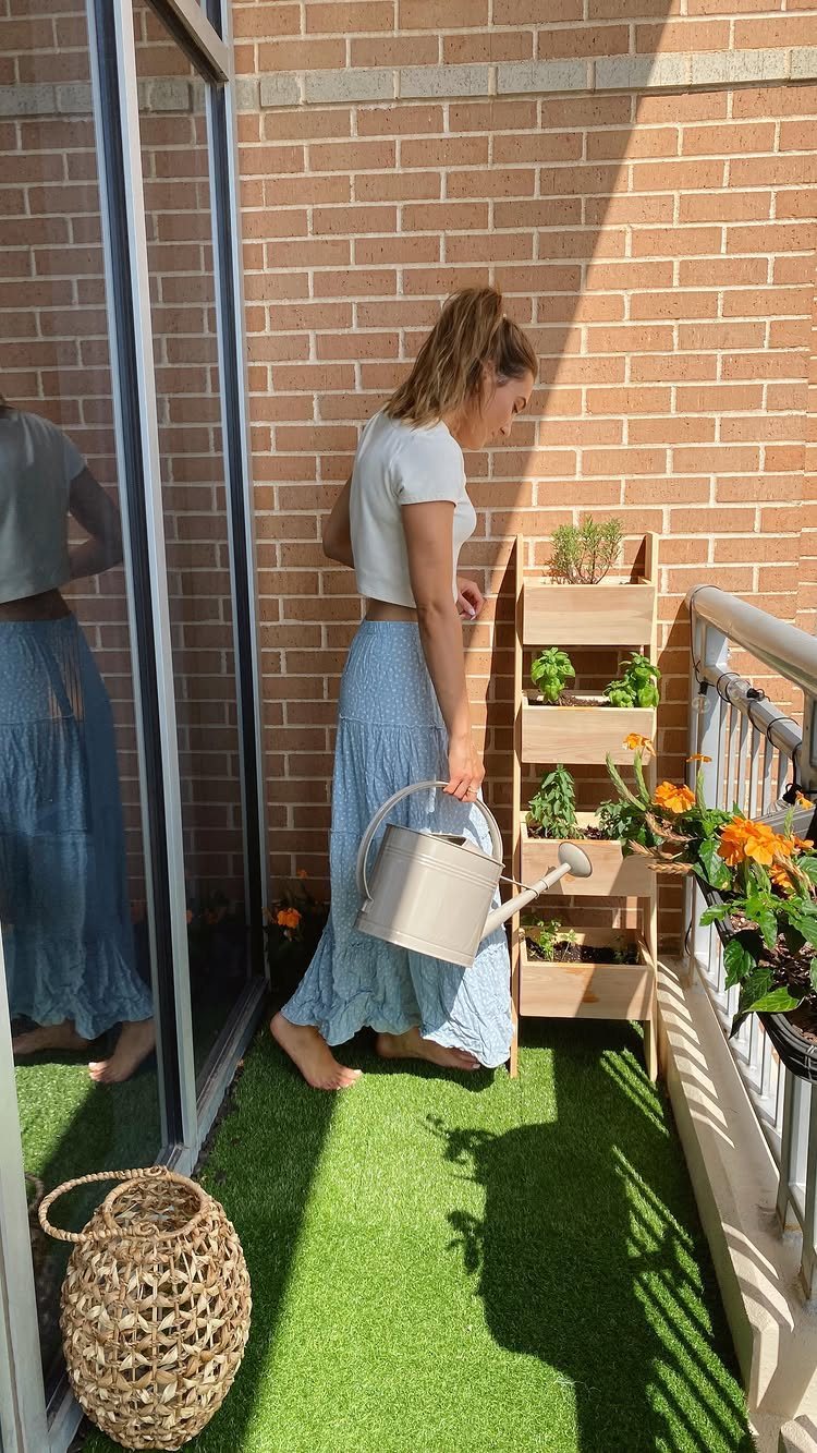 12 Container Garden Ideas for Small Patios, Balconies & Rooftops 12 A woman holding a watering can next to a wooden tiered plant stand on a balcony.