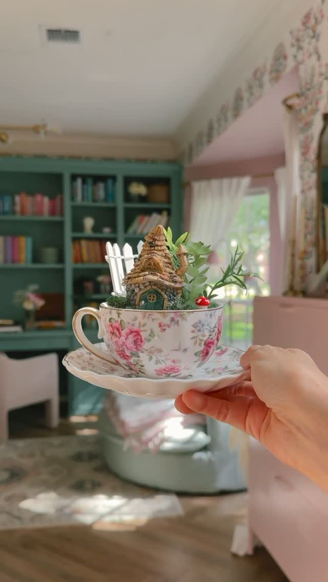 13 Fairy Garden Ideas: Broken Pots to Teacup Worlds 13 A hand holding a floral teacup containing a miniature fairy garden with a tiny house.