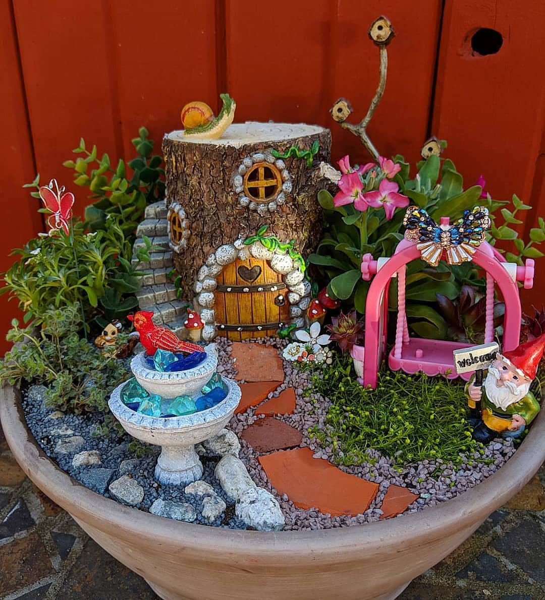 13 Fairy Garden Ideas: Broken Pots to Teacup Worlds 10 A miniature fairy garden in a planter with a tree stump house and pink swing.