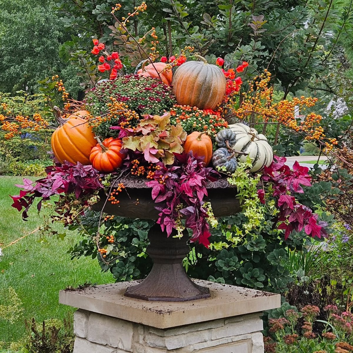 Fall Planter Ideas: 12 Container Combos for Color, Foliage and Texture 6 A dark metal urn filled with pumpkins, colorful autumn leaves, and bright berries on a stone pedestal.