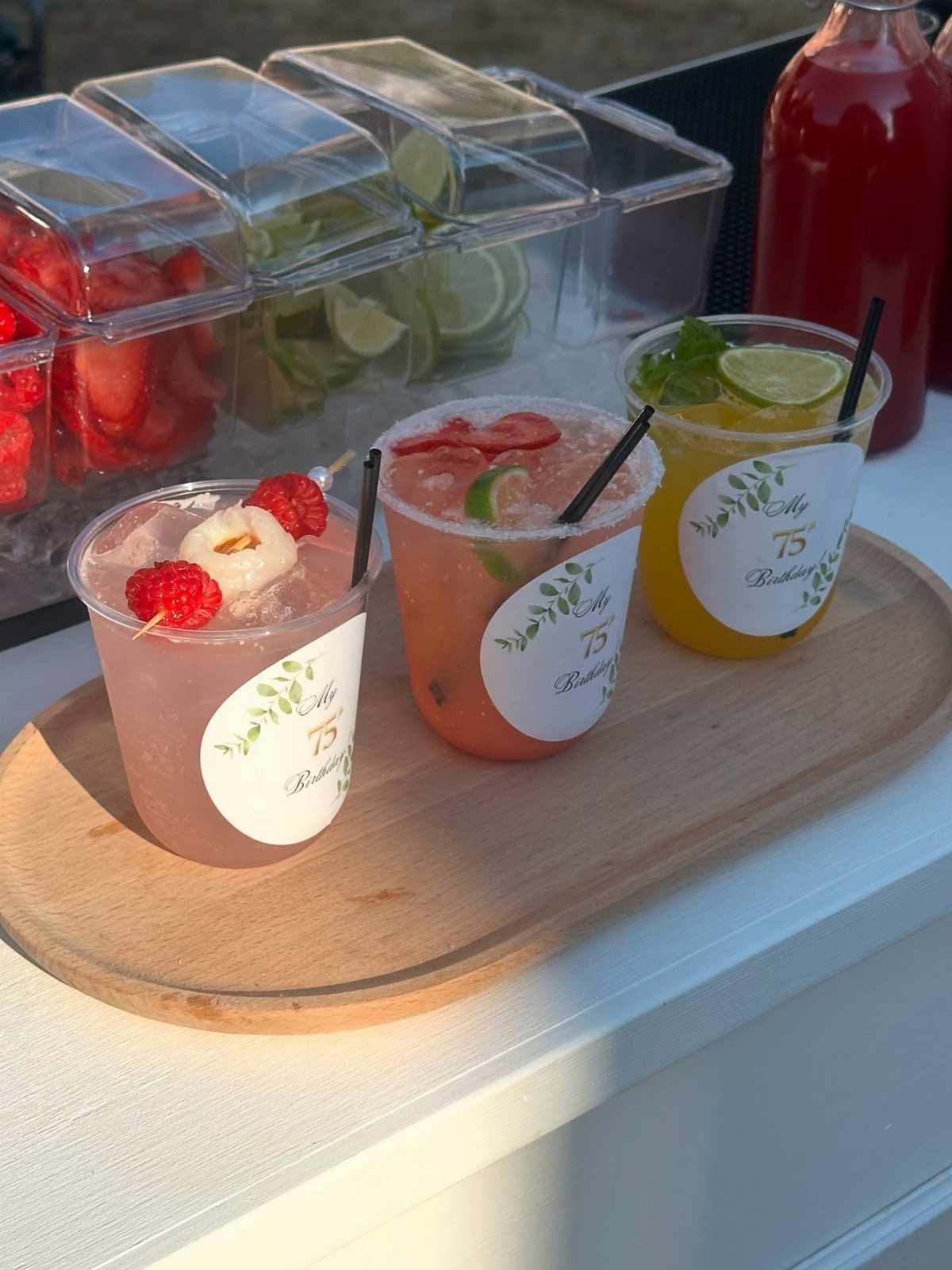 Three colorful cocktails with customized white labels sit on a wooden serving board.