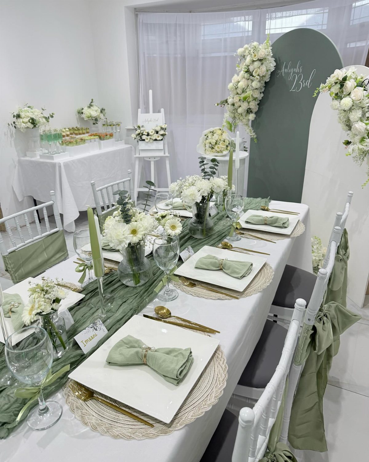 A long dining table decorated with sage green linens, white flowers, and gold cutlery.