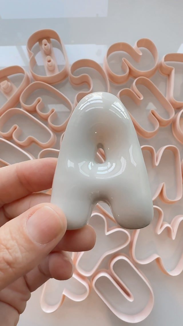 A hand holding a glossy white 3D bubble letter A against pink cookie cutters.