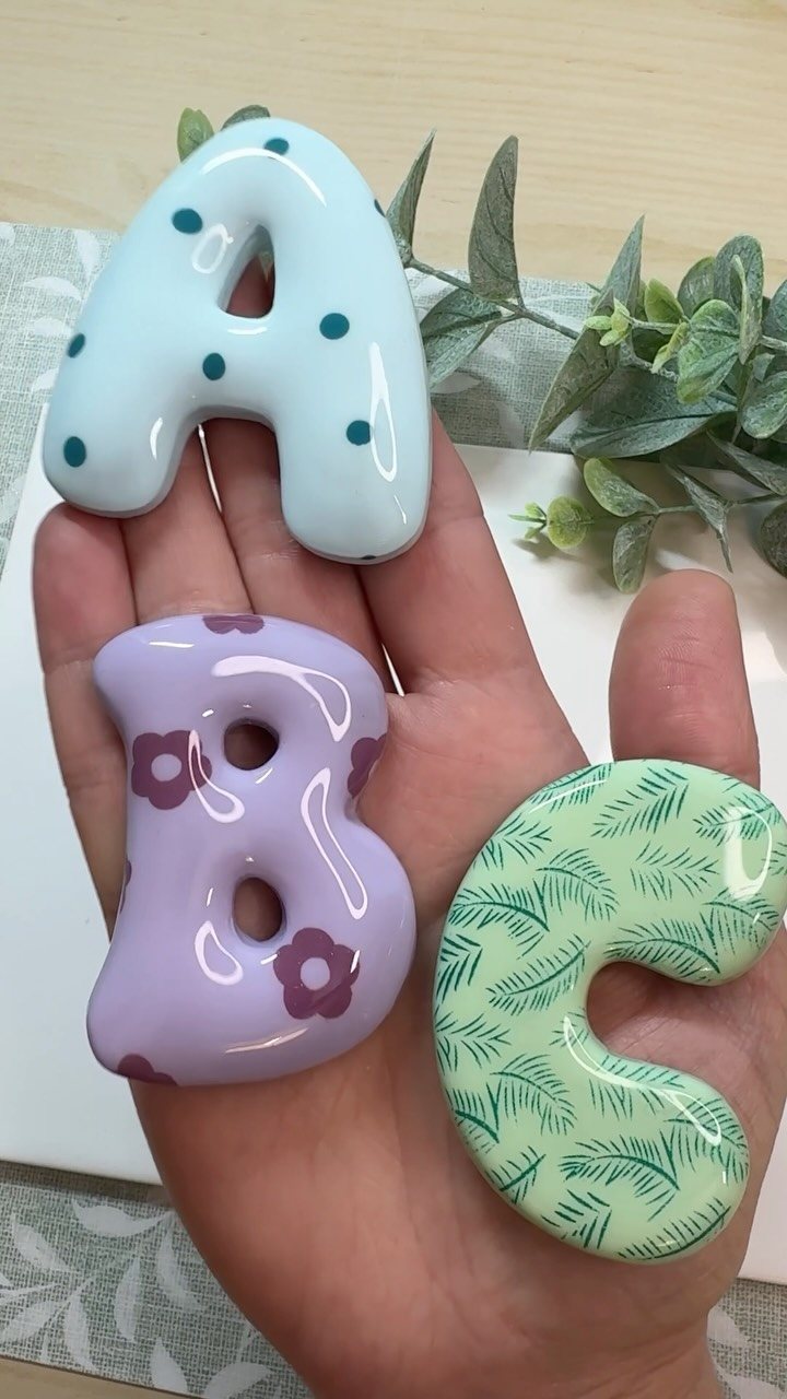A hand holding three decorated, glossy bubble letters A, B, and C with various patterns.