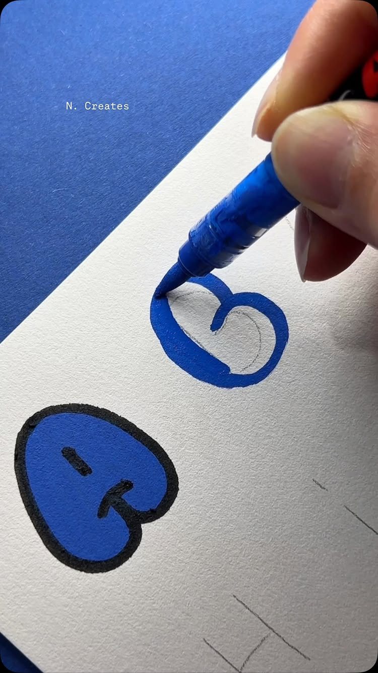 A hand drawing a blue bubble letter B on white paper with a blue marker.