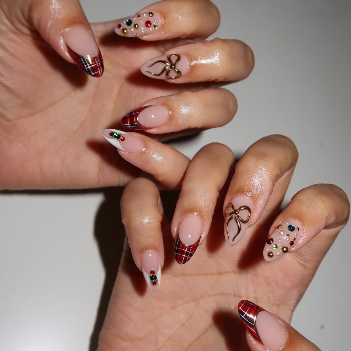 22 Christmas Nail Designs Ideas for 2026 Holiday Manicures 8 Manicured hands showing festive nails with plaid, gold bows, and colorful rhinestones.