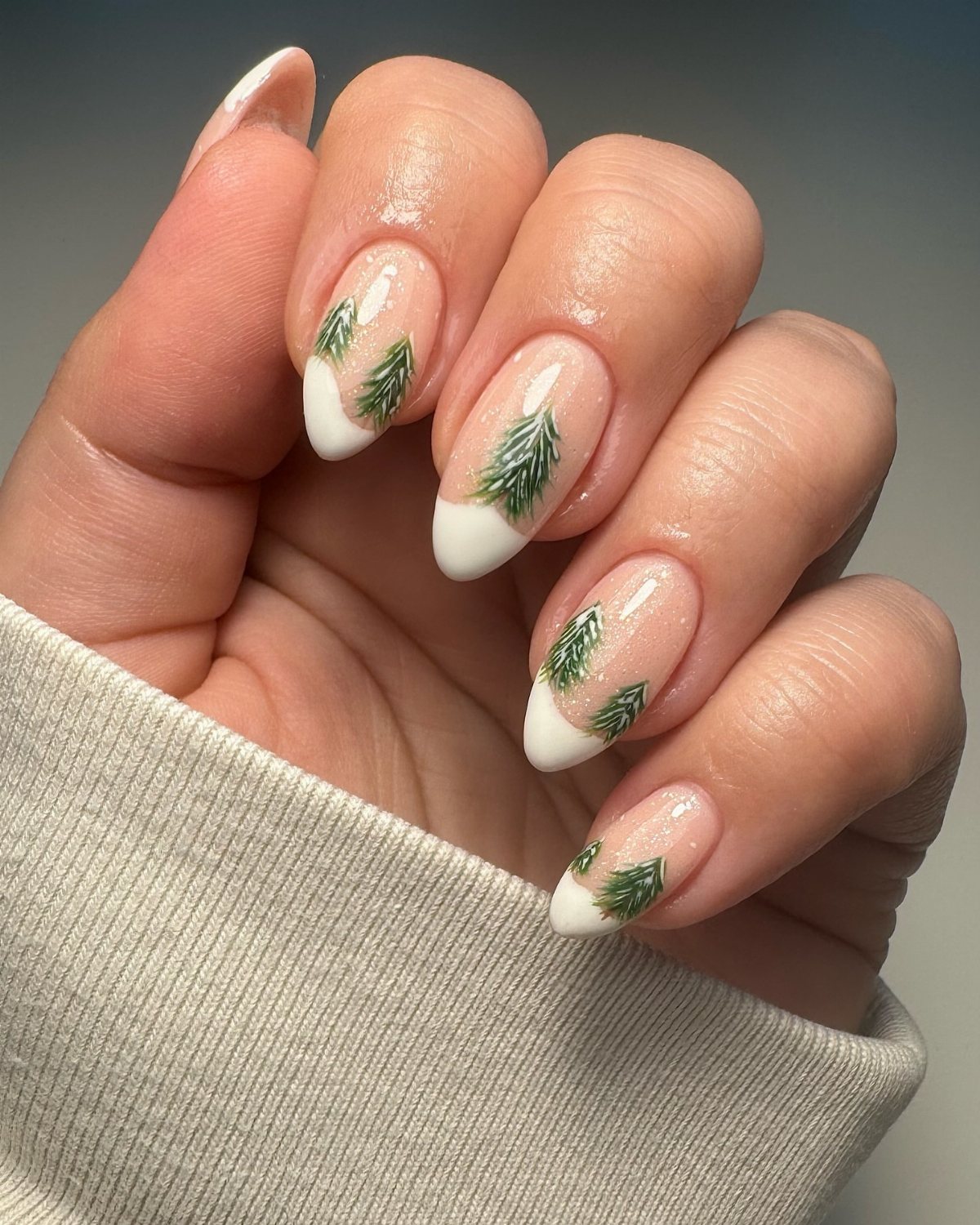 22 Christmas Nail Designs Ideas for 2026 Holiday Manicures 7 Hand with almond-shaped nails featuring white tips and small green Christmas tree designs.