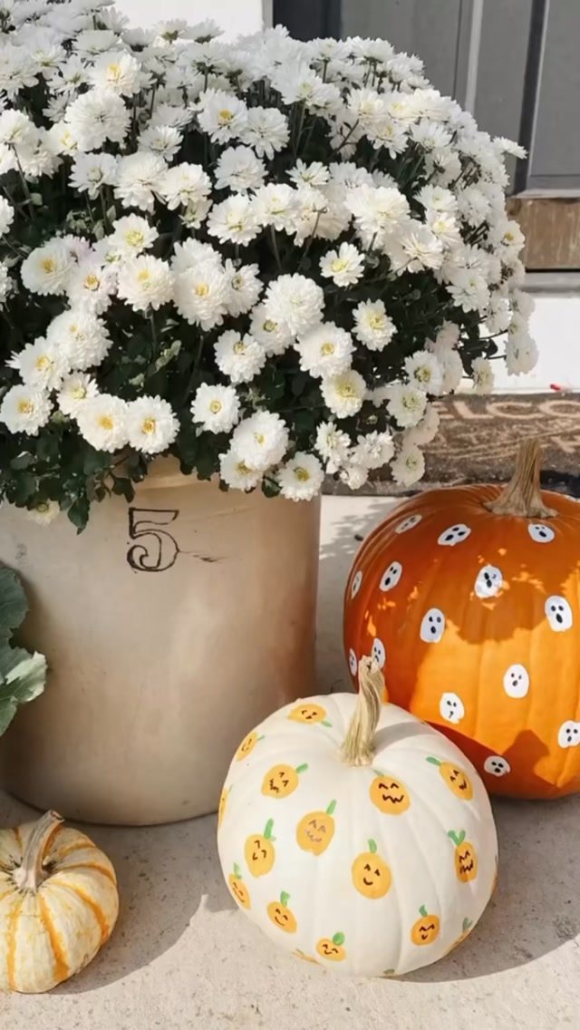 Painted pumpkins with ghost and pumpkin patterns sitting next to white chrysanthemums.
