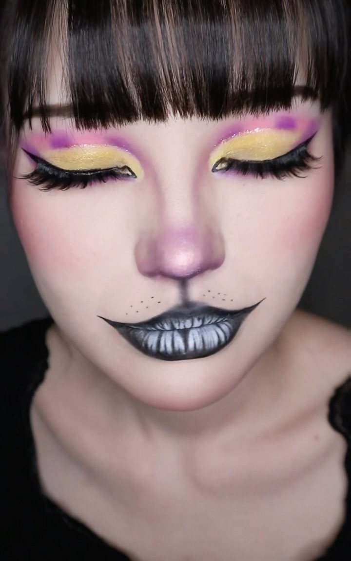 Person wearing colorful cat makeup with yellow eyeshadow, pink nose, and black lips with teeth.