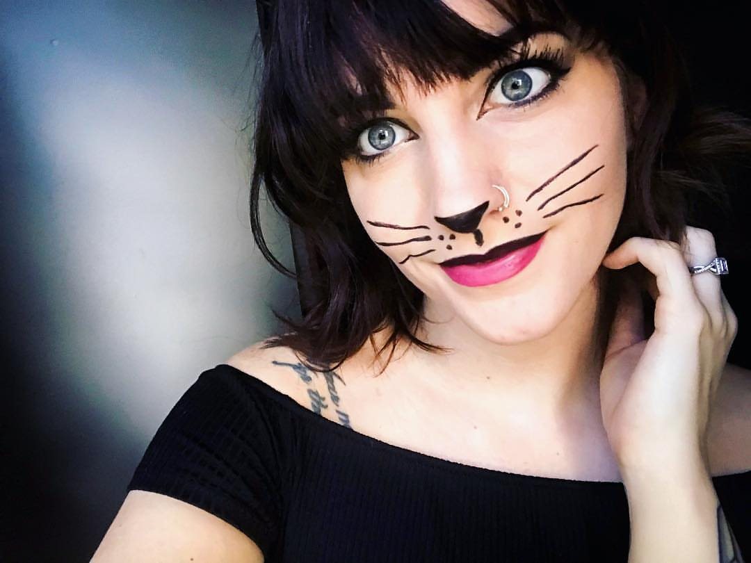 Woman with dark hair wearing black cat whiskers and nose face paint.
