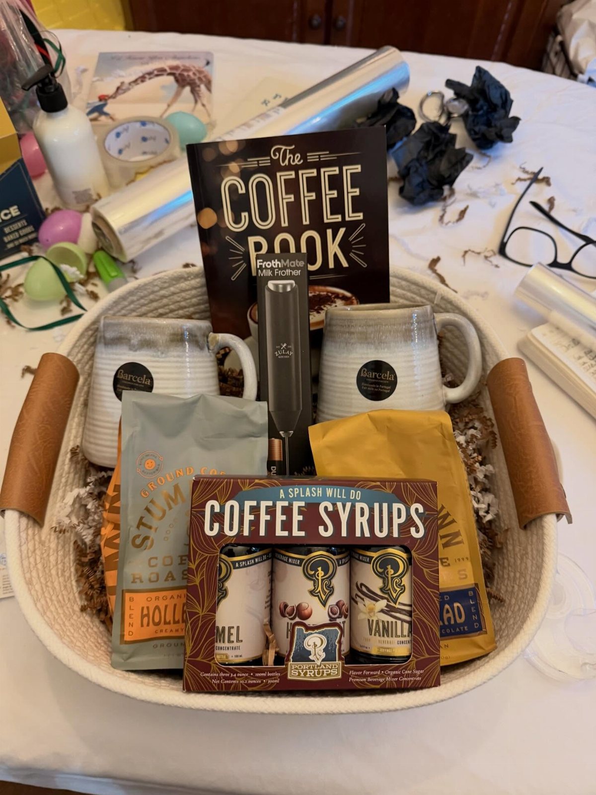 A gift basket filled with coffee mugs, coffee syrups, coffee beans, and a coffee book.