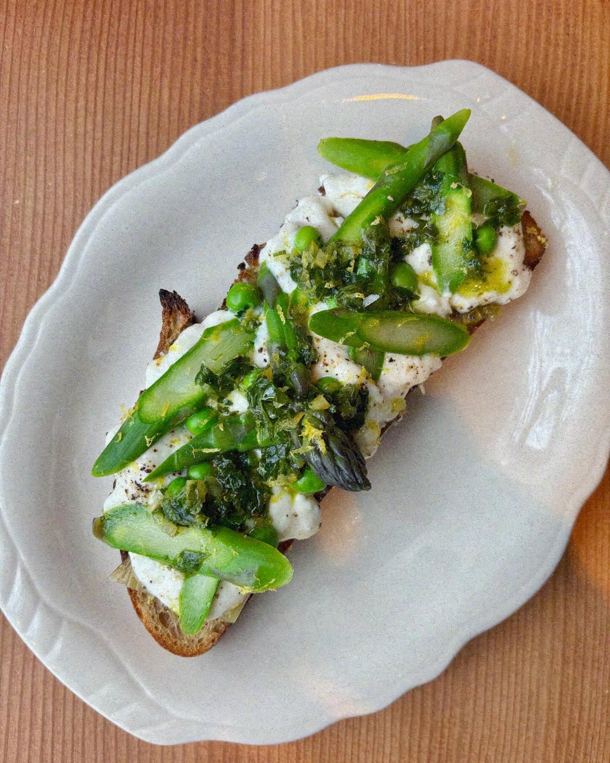 Toasted bread topped with white cheese, green asparagus, peas, and pesto on a white plate.