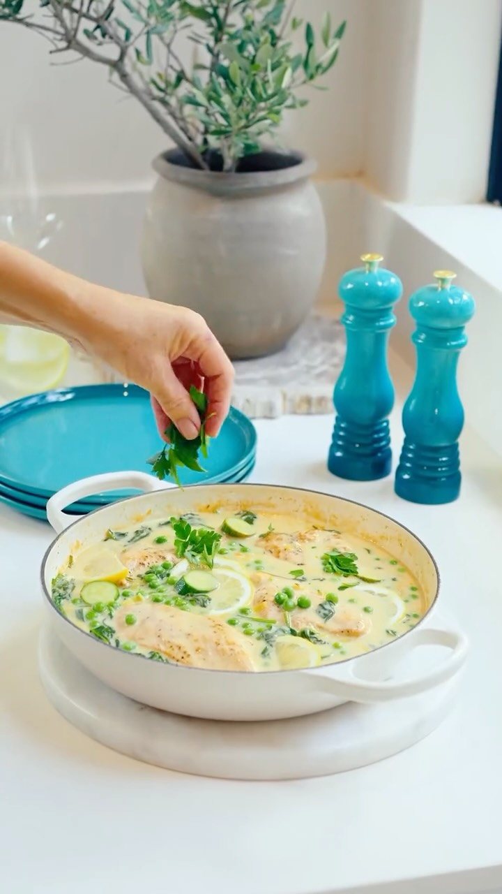 A hand garnishes a creamy chicken dish with fresh green herbs in a white pan.