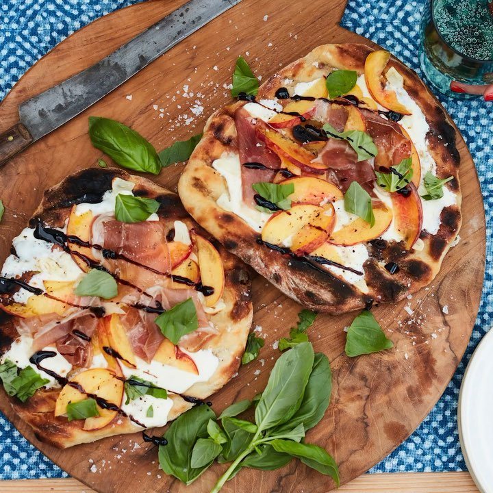 Two rustic peach and prosciutto pizzas topped with basil on a wooden board.