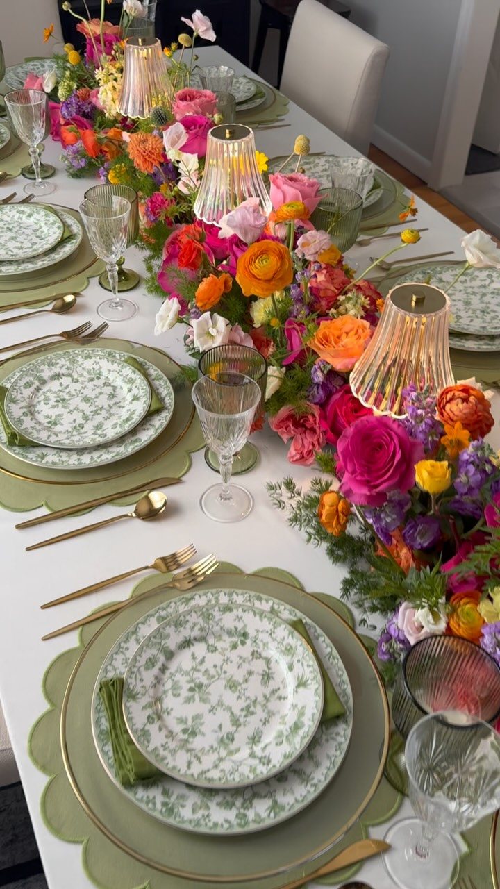 A long dining table decorated with a vibrant floral runner, green patterned plates, and gold cutlery.