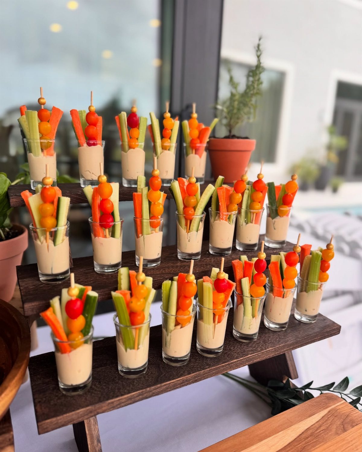 Vegetable skewers with dip in small glasses arranged on a tiered wooden serving stand.