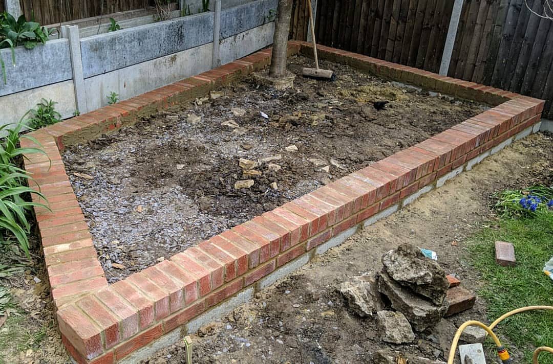 18 Front Raised Flower Bed Ideas, From Cottage Beds to Cinder Blocks 3 A rectangular red brick raised garden bed containing dark soil and a single tree trunk.