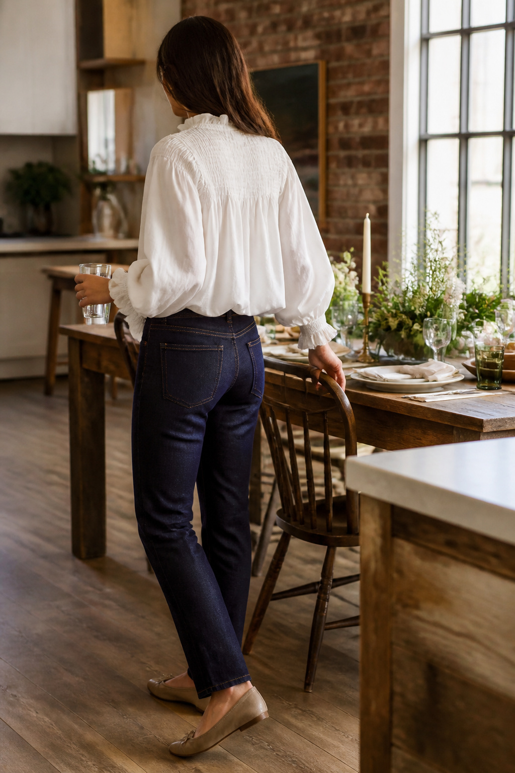 A model wearing jeans and a white blouse in a relaxed kitchen.