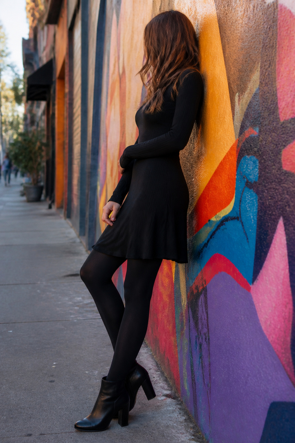 A model in a fitted black turtleneck dress and black tights stands by a mural.