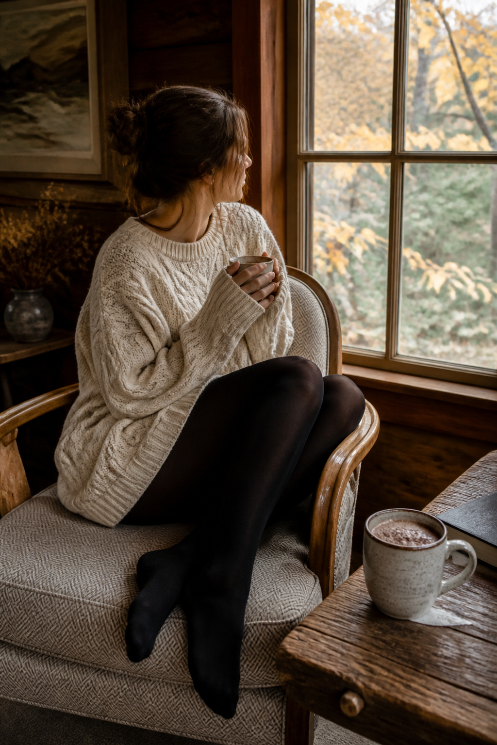 A model in a cream sweater and black tights sits by a window in a cabin.