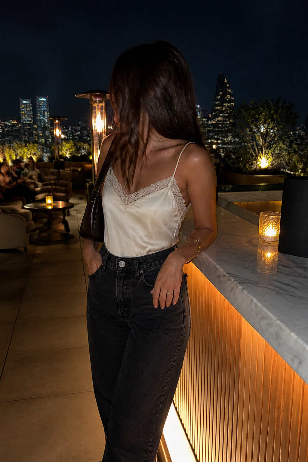 A woman in a lace-trim top and dark jeans at a rooftop bar.