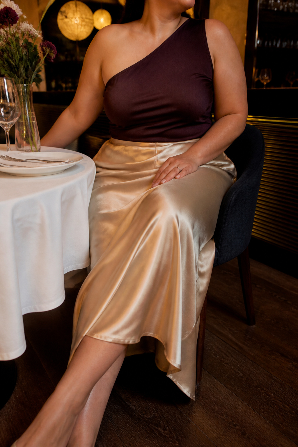 One-shoulder top paired with a satin skirt in a dimly lit restaurant.